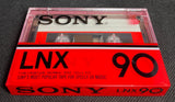 A vintage Sony 1978 LNX 90 cassette tape and its original red-and-black plastic case, displayed on a dark textured surface.