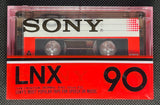 A vintage Sony 1978 LNX 90 cassette tape and its original red-and-black plastic case, displayed on a dark textured surface.